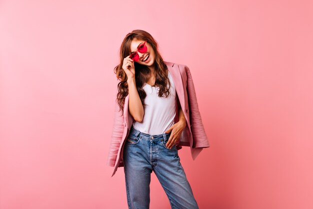 Optimistische Ingwerdame in der hellen Sonnenbrille lachend. Lächelndes atemberaubendes Mädchen in den Jeans, die auf Rosa aufwerfen.