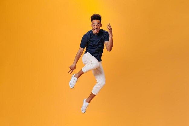 Optimistischer dunkelhäutiger Typ in stylischem Outfit, der auf orangefarbenem Hintergrund springt Brünetter Mann in schwarzem T-Shirt und weißer Hose posiert isoliert