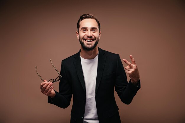 Optimistischer Mann in schwarzer Jacke hält Brille und zeigt Friedenszeichen