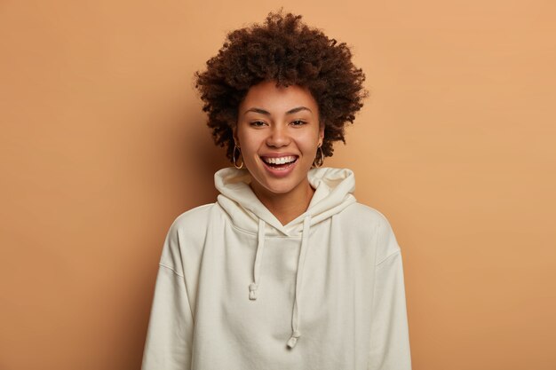 Optimistisches junges Mädchen in lässigem weißem Sweatshirt, lächelt glücklich, steht gegen braunen Raum, hört lustigen Witz von Freund