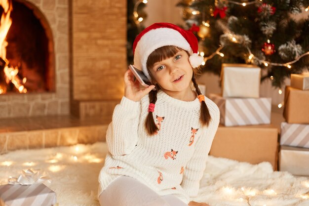 Optimistisches kleines Mädchen mit weißem Pullover und Weihnachtsmann-Hut, Blick in die Kamera, festliche Stimmung, Telefonieren, Sitzen auf dem Boden in der Nähe von Weihnachtsbaum, Geschenkboxen und Kamin.