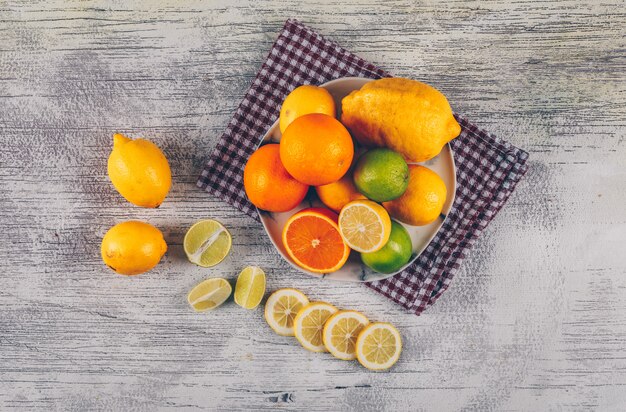 Orange mit grünen und gelben Zitronen mit Scheiben in einem Teller auf Stoff und grauem hölzernem Hintergrund, Draufsicht.