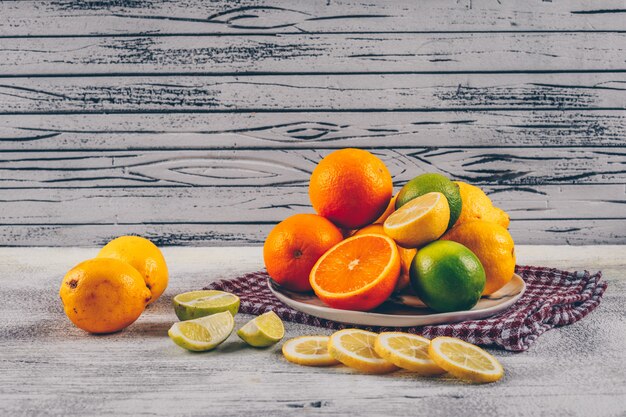 Orange mit grünen und gelben Zitronen mit Scheiben in einem Teller und auf Stoff und grauem hölzernem Hintergrund, Seitenansicht.