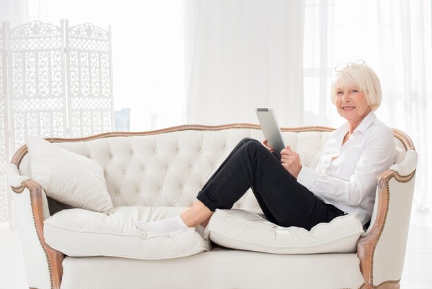 Ordentliche alte Frau, die auf Sofa mit einer Tablette sitzt
