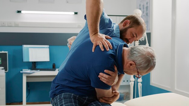Osteopath hilft Senioren, Knochen im Kabinett zu knacken, und macht Physiotherapieübungen. Männlicher Assistent, der ein physiotherapeutisches Verfahren verwendet, um die Mobilität zu erhöhen und den Patienten zu behandeln.