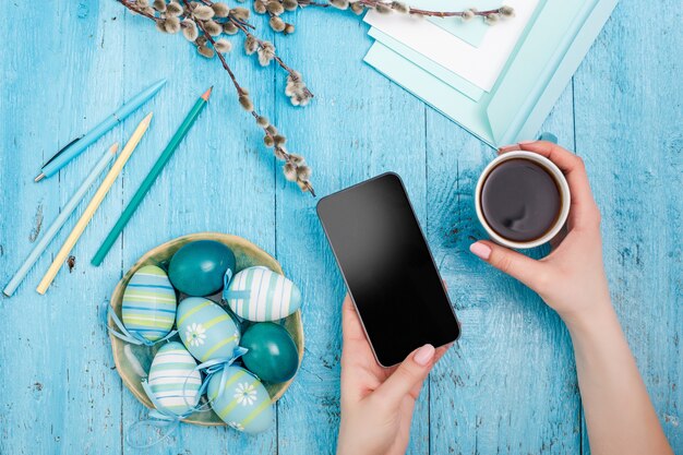 Ostern im Büroarbeitsplatz auf blauem Holztisch. weibliche Hände mit Telefon und einer Tasse Kaffee