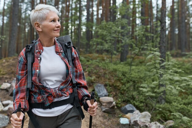 Outddor Porträt der glücklichen europäischen Rentnerin mit Rucksack und Stangen, schöne Natur genießend, während Nordic Walking im Kiefernwald.