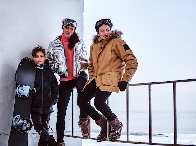 Outdoor-Aktivitäten in den Winterferien. Familie in stylischer Kleidung posiert mit Snowboard neben einem Geländer am verschneiten Strand