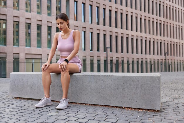 Outdoor-Aufnahme einer aktiven sportlichen Frau, die sich in einer städtischen Umgebung ausruht, atmet tief ein, nachdem das Training Sportkleidung trägt und Turnschuhe müde sind