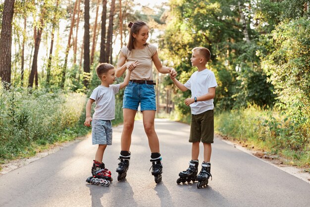 Outdoor-Aufnahme einer glücklichen Familie, die im Sommerpark Spaß und Rollschuhlaufen hat, Mama, die Kinderhändchen hält, froh ist, das Wochenende zusammen zu verbringen, aktiver Zeitvertreib.