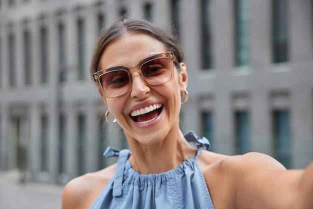 Outdoor-Aufnahme einer glücklichen jungen Frau, die im Allgemeinen lächelt, trägt eine trendige Sonnenbrille und macht Selfie, während sie im Freien in der Stadt spaziert