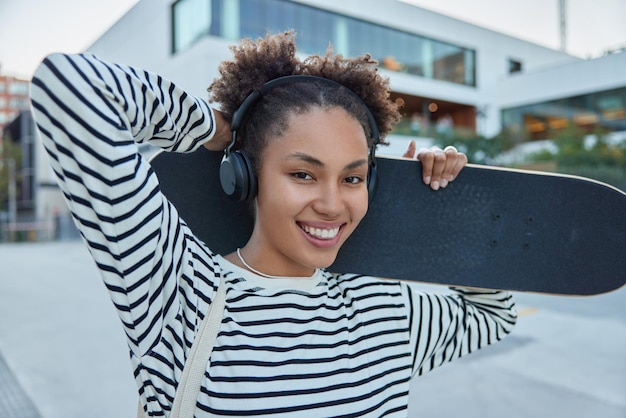 Outdoor-Aufnahme einer positiven Skaterin genießt die Freizeit für ihr Lieblingshobby hält Skateboard und hört Musik in drahtlosen Kopfhörern trägt gestreifte Jumper-Posen gegen die verschwommene Stadt