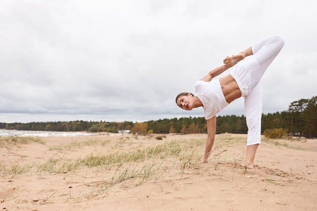 Outdoor-Bild in voller Länge von einer konzentrierten kaukasischen Frau, die ein weißes Outfit trägt, Yoga im Freien macht, mit einem Fuß und einer Hand auf Sand steht, Balance, Konzentration und Koordination trainiert