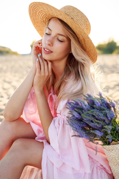 Outdoor-Lebensstilporträt der anmutigen weißen Frau, die am sonnigen Strand nahe Ozean sitzt. Strohhut tragen. Naturhintergrund.