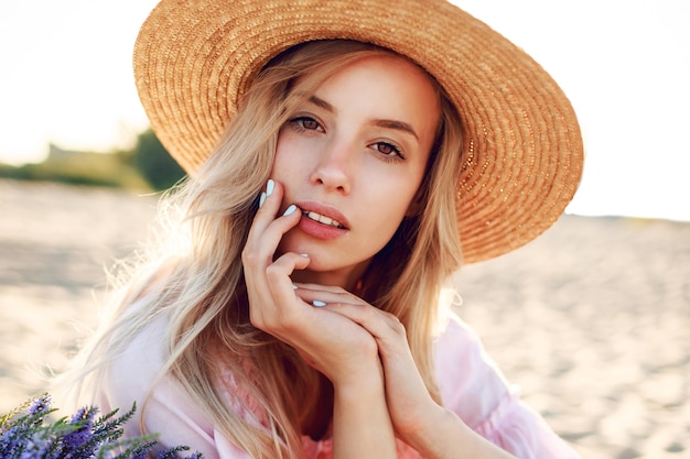 Outdoor-Lebensstilporträt der anmutigen weißen Frau, die auf sonnigem Strand nahe Ozean steht. Strohhut tragen. Naturhintergrund. Warme weiche Farben.