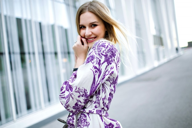 Outdoor-Lifestyle-Porträt der verlassenen lächelnden blonden Frau, die elegantes trendiges Blumenkleid trägt und in der Kamera schaut, Frühlingssommerzeit.