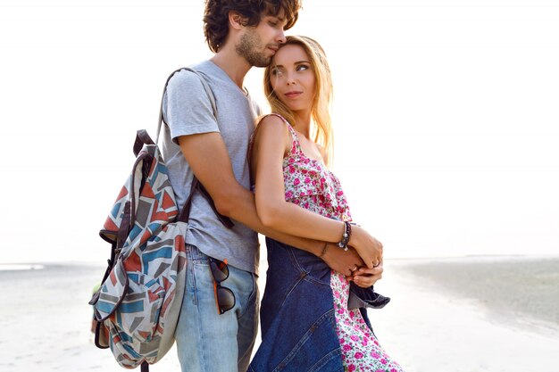 Outdoor-Lifestyle-Porträt des erstaunlichen hübschen jungen Paares in der Liebe, die am Strand aufwirft. Stilvoller Mann und Frau umarmen sich und verbringen viel Zeit miteinander. Blumenkleid Rucksack und Jeans.