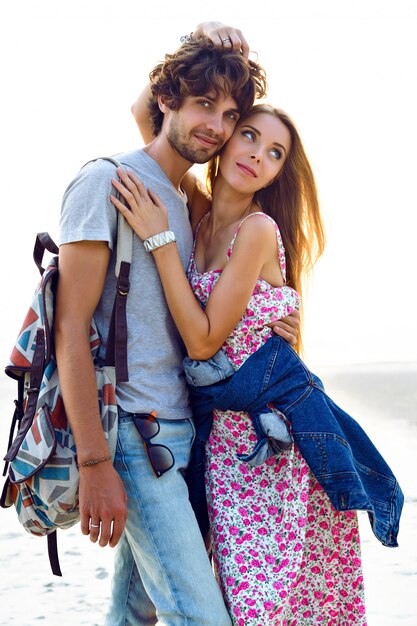 Outdoor-Lifestyle-Porträt des erstaunlichen hübschen jungen Paares in der Liebe, die am Strand aufwirft. Stilvoller Mann und Frau umarmen sich und verbringen viel Zeit miteinander. Blumenkleid Rucksack und Jeans.
