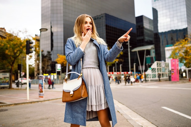 Outdoor-Lifestyle-Porträt einer blonden hübschen jungen Geschäftsfrau, die in einem modernen Gebäudebereich spaziert, einen blauen Mantel und ein weibliches graues Kleid trägt, überraschte unheimliche Gefühle und zeigte etwas durch ihren Finger.