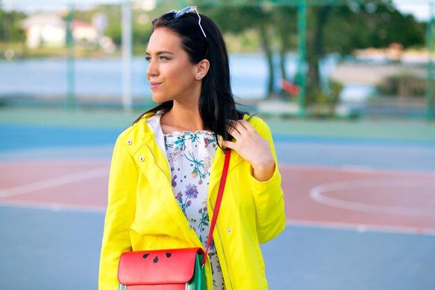 Outdoor-Lifestyle-Porträt einer brünetten Frau mit gelbem Regenmantel und stilvoller Tasche, die auf dem Sportplatz posiert, Frühlingsherbstsaison.