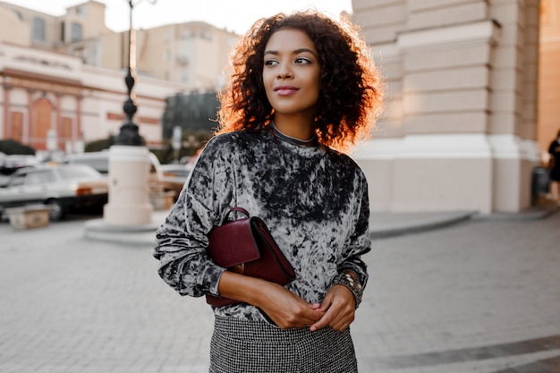 Kostenloses Foto outdoor-mode-porträt der glamourösen sinnlichen jungen stilvollen schwarzen dame, die trendiges herbstoutfit, grauen samtpullover und luxusgeldbörse trägt.