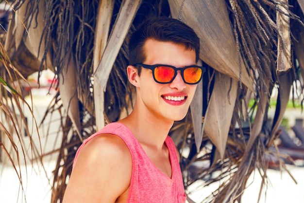 Outdoor-mode-porträt des schönen stilvollen kerls, der große zeit am tropischen strand verbringt, nahe kokospalme aufwirft, helles outfit und neon-sonnenbrille trägt.