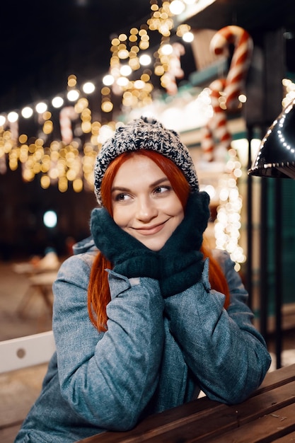 Outdoor-Nachtfoto eines jungen, schönen, glücklich lächelnden Mädchens, das festliche Dekoration genießt, auf dem Weihnachtsmarkt posiert, in der Straße der europäischen Stadt, mit gestrickter Beanie-Mütze