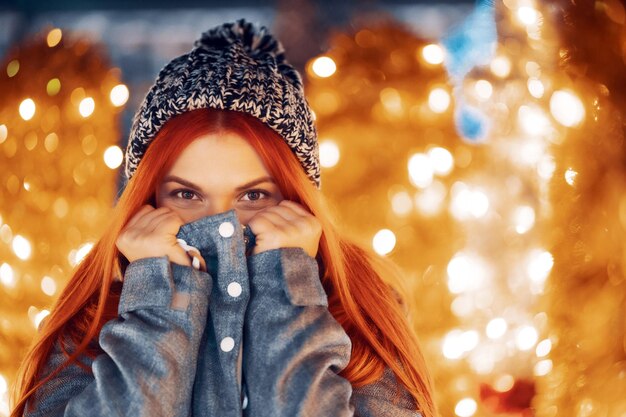 Outdoor-Nachtfoto eines jungen, schönen, glücklich lächelnden Mädchens, das festliche Dekoration genießt, auf dem Weihnachtsmarkt posiert, in der Straße der europäischen Stadt, mit gestrickter Beanie-Mütze