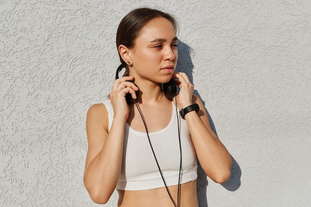 Outdoor-Porträt einer jungen erwachsenen schönen Frau, die weißes Top trägt, während des Trainings Musik hört, die Hände an den Kopfhörern hält und mit nachdenklichem Gesichtsausdruck wegschaut.