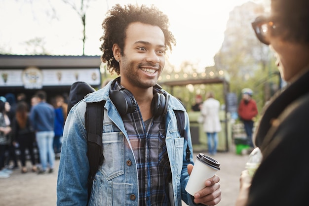 Outdoor-Porträt eines stilvollen, fröhlichen afroamerikanischen Mannes mit Borsten und Afro-Frisur, der eine Tasse Kaffee hält und breit lächelt, während er mit einer attraktiven Frau im Park spricht