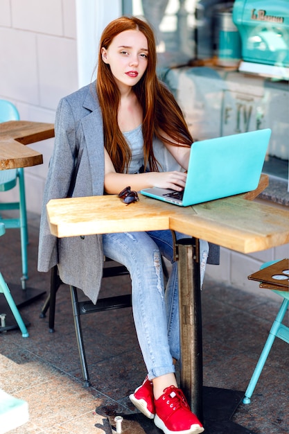 Outdoor-Stadtmodeporträt der jungen Geschäftsfrau, die am Café auf der Terrasse am sonnigen Tag arbeitet, lässiges stilvolles Outfit, Minzdetails, unter Verwendung ihres Laptops, Cafépause, Geschäftskonzept.