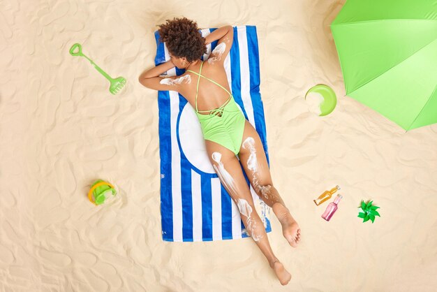 Overhead-Aufnahme einer entspannten, schlanken Frau in grünem Badeanzug liegt auf dem Bauch, sonnt sich am Strand und trägt Hautpflegecreme auf