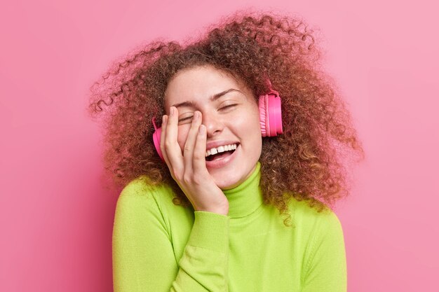 Overyoyed Teenager-Mädchen mit lockigen, buschigen Haaren lächelt breit hält die Hand auf dem Gesicht schließt die Augen vor Vergnügen hört Musik über drahtlose Kopfhörer, die lässig isoliert auf rosa Wand gekleidet sind