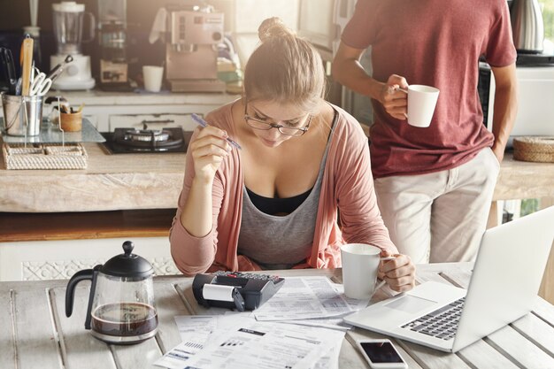 Paar, das gemeinsam das Inlandsbudget verwaltet. Junge Frau in Gläsern, die Stift halten, während sie Berechnungen unter Verwendung des Taschenrechners und des Laptops machen