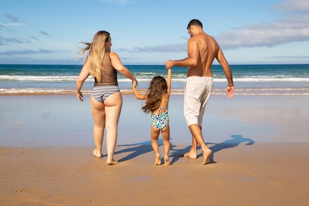 Paar Eltern und kleine Tochter, die Badeanzüge tragen und auf goldenem Sand zum Wasser gehen