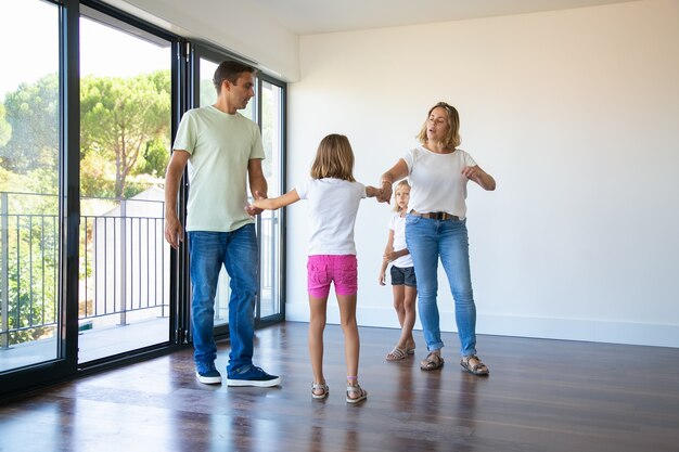 Paar Eltern und zwei Kinder genießen ihr neues Zuhause, stehen in einem leeren Raum und halten Hände, tanzen