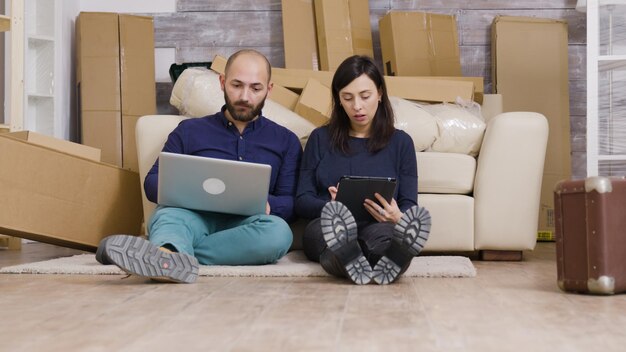 Paar in ihrer neuen wohnung, die auf dem boden sitzt und online möbel mit laptop und tablet kauft.