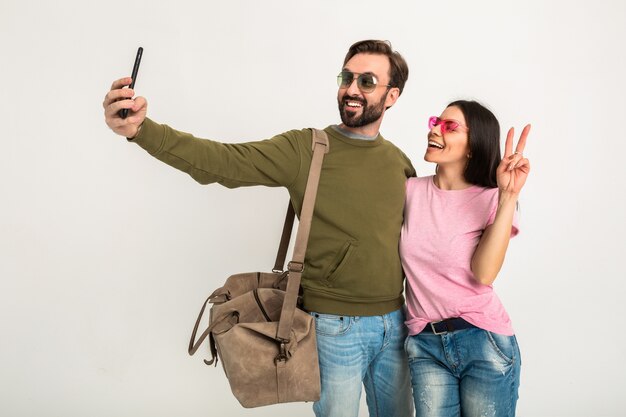 Paar isoliert, hübsche lächelnde Frau im rosa T-Shirt und Mann im Sweatshirt mit Reisetasche, Jeans und Sonnenbrille tragend, Spaß haben, zusammen reisen und lustiges Selfie-Foto am Telefon machen