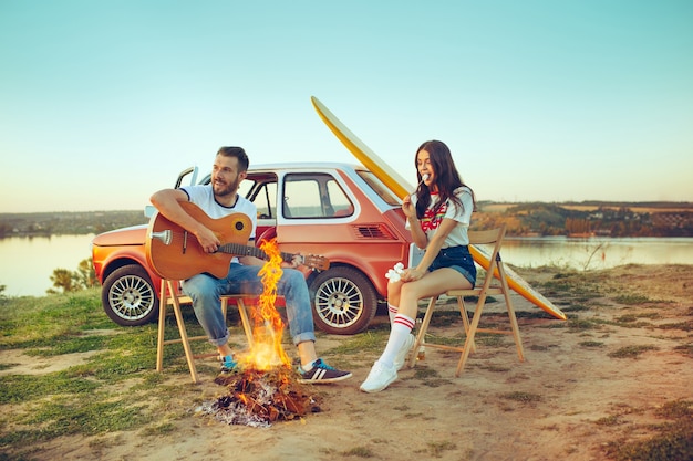 Paar sitzt und ruht sich am Strand aus und spielt Gitarre an einem Sommertag in der Nähe des Flusses