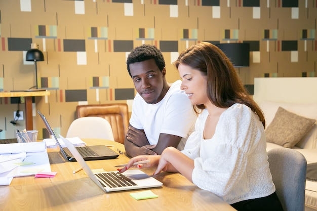 Paar verschiedene Designer, die mit Laptops und Blaupausen am Tisch sitzen und Designprojekt diskutieren