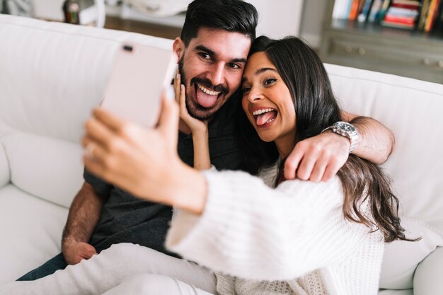 Paare, die auf dem Sofa zeigt ihre Zunge heraus nimmt selfie auf Smartphone sitzen
