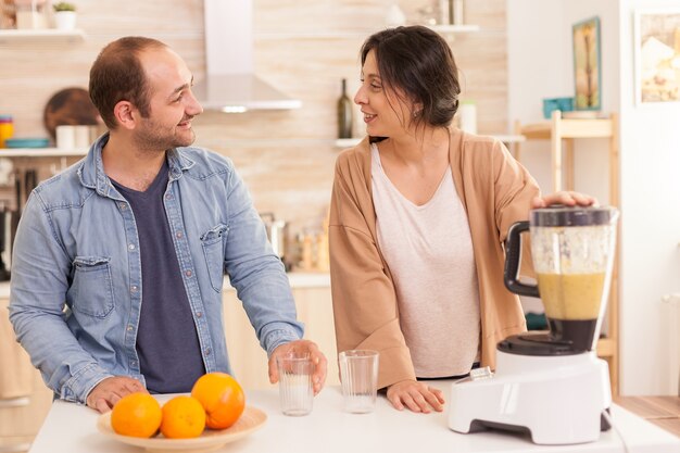 Paare, die einander anlächeln, während sie nahrhafte Früchte in der Küche mit Mixer zubereiten. Gesunder, unbeschwerter und fröhlicher Lebensstil, Ernährung und Frühstückszubereitung am gemütlichen sonnigen Morgen