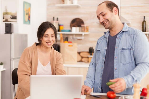 Paare, die Online-Rezept auf Laptop in der Küche für Gemüsesalat ansehen. Mann hilft der Frau, gesundes Bio-Abendessen zuzubereiten und zusammen zu kochen. Romantisch fröhliche Liebesbeziehung