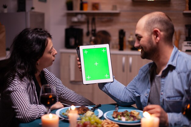 Paare, die Tablet-PC mit Chroma-Key verwenden und ihre gemeinsame Zeit beim romantischen Abendessen genießen. Mann und Frau betrachten die Chroma-Key-Anzeige der Greenscreen-Vorlage, die am Tisch in der Küche sitzt