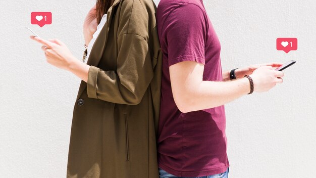 Paare, die zurück zu Rückseite unter Verwendung des Mobiltelefons mit Liebesmitteilungsikonen gegen Wand stehen