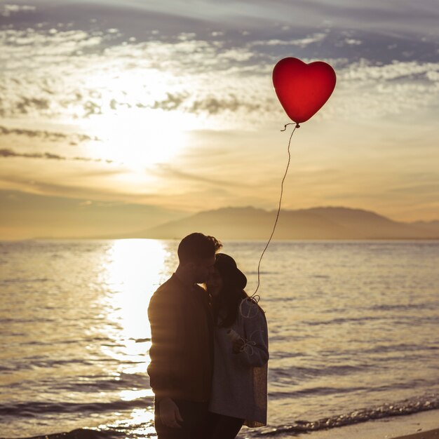 Paare mit dem Herzballon, der auf Seeufer am Abend umarmt