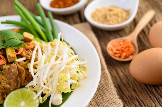 Pad Thai in einem weißen Teller mit Zitrone, Eiern und Gewürzen auf einem Holztisch.