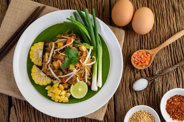 Pad Thai in einem weißen Teller mit Zitrone, Eiern und Gewürzen auf einem Holztisch.