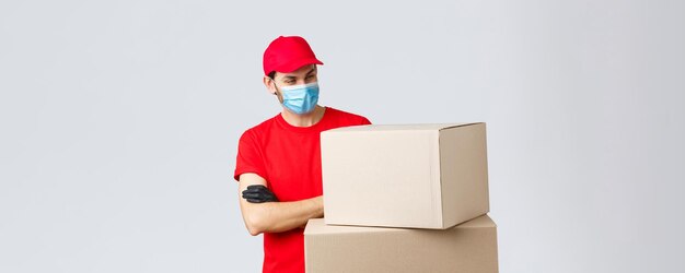 Paket- und Paketzustellung covid19 Quarantäne- und Überweisungsaufträge Neugieriger männlicher Kurier in roter einheitlicher medizinischer Gesichtsmaske und Handschuhen, die interessiert auf Kisten mit gekreuzten Armen blicken, starren fasziniert