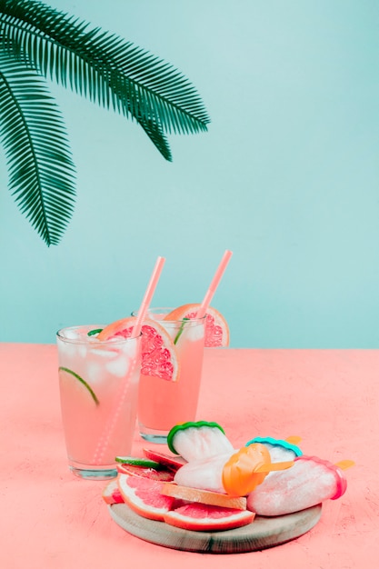 Kostenloses Foto palmblätter über den grapefruit-cocktailgläsern; eis am stiel auf korallenschreibtisch gegen aquamarinen hintergrund